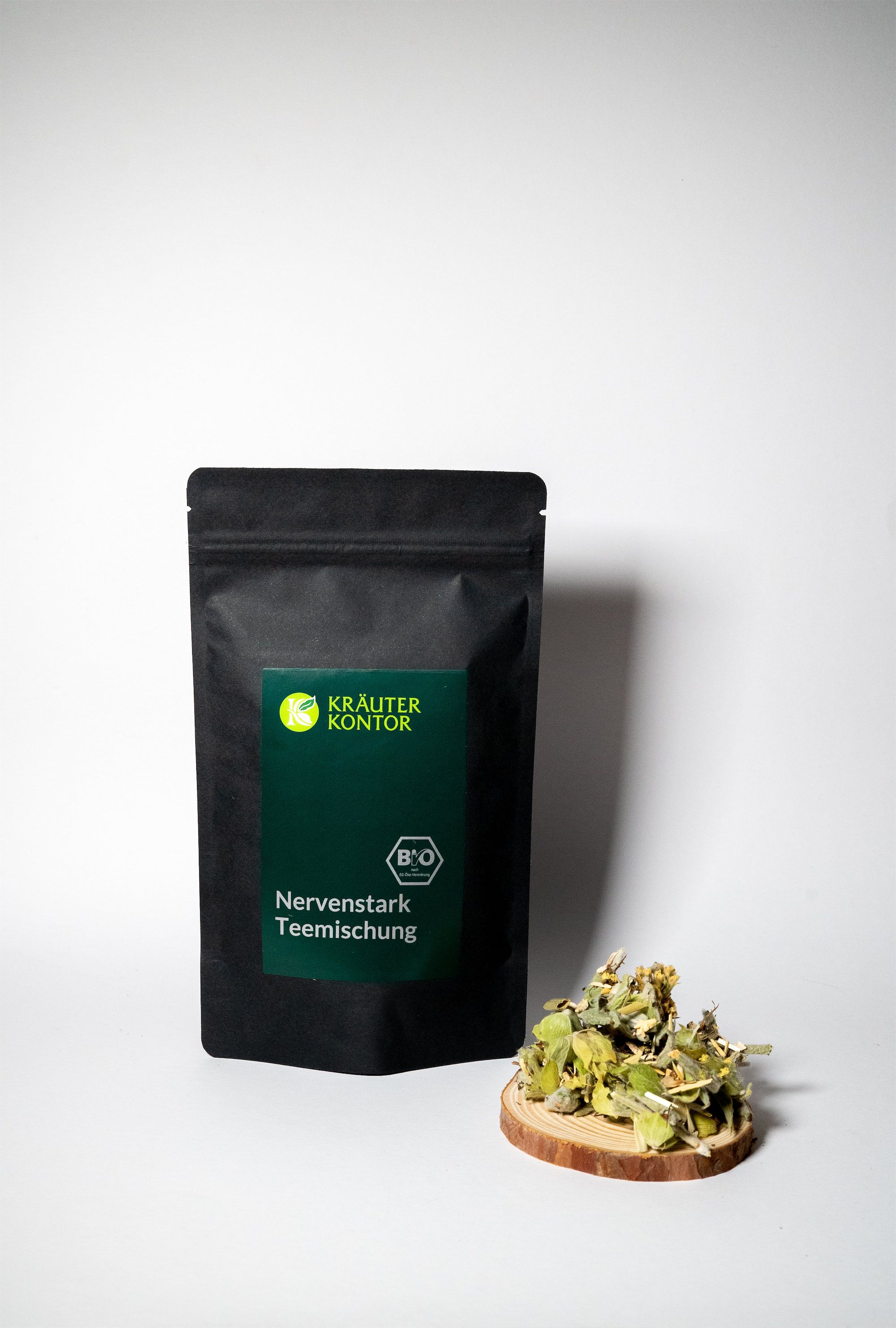 Schwarzer Beutel mit grünem Etikett. Text: Kräuterkontor, Nervenstark Teemischung. Bio-Siegel. Kräuter auf Holzscheibe.