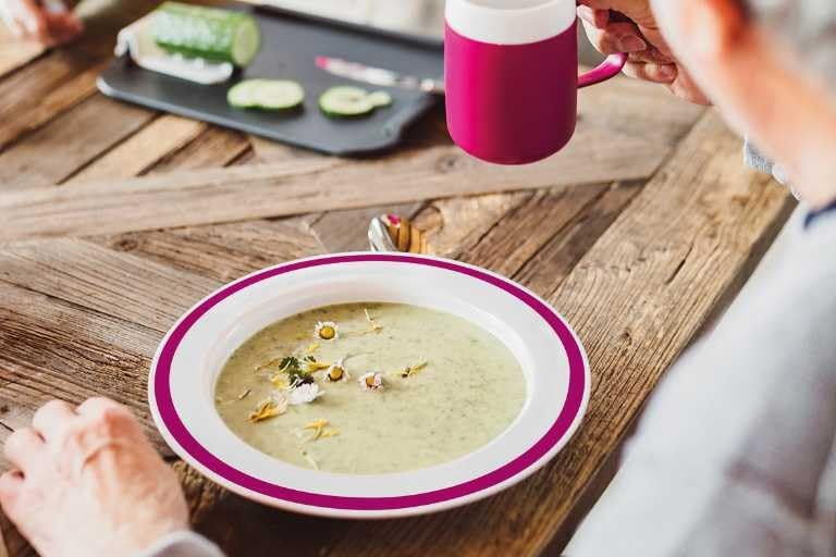 Teller mit Suppe. Weiß mit lila Rand. Person hält Tasse. Holz-Tisch.