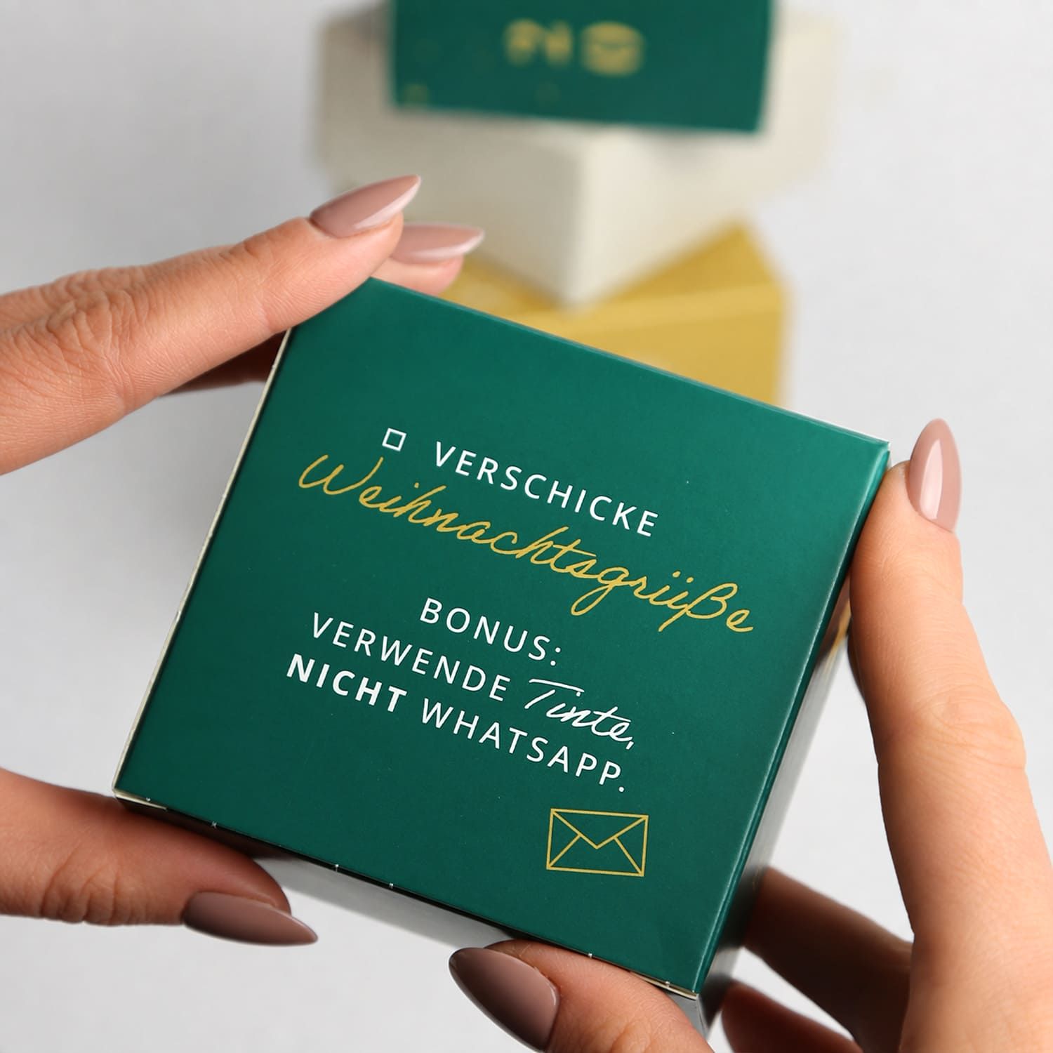 Eine Hand hält eine kleine grüne Schachtel mit goldenem Text: 'Verschicke Weihnachtsgruße'.