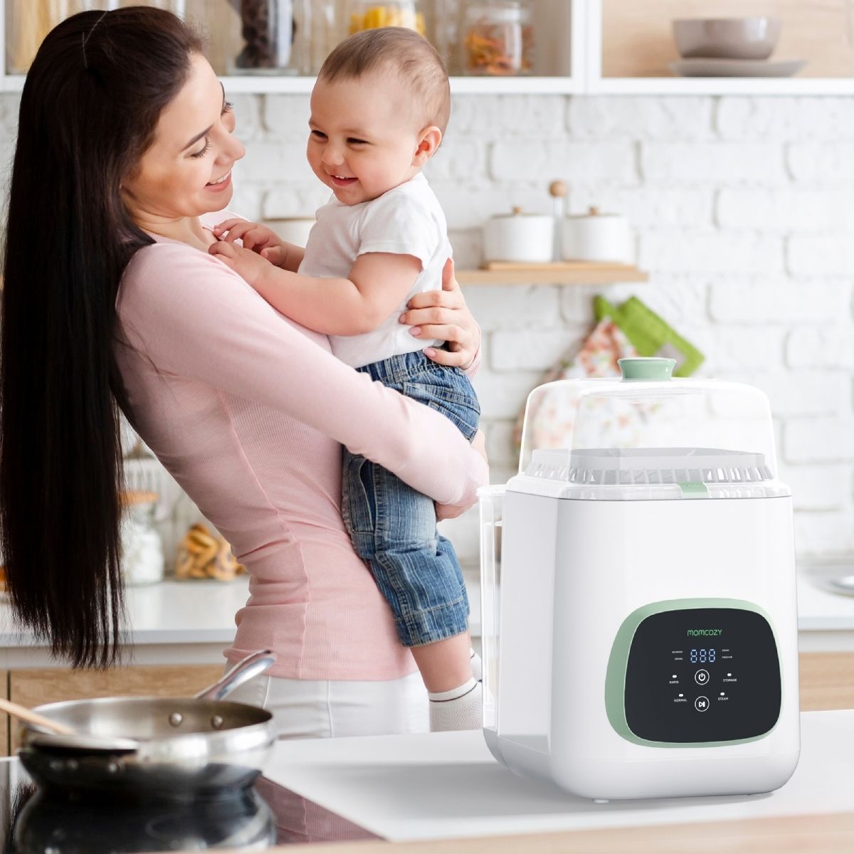 Frau hält Baby vor Babyflaschenreiniger. Reiniger weiß mit grünem Akzent. Momcozy-Logo sichtbar. Küche im Hintergrund.