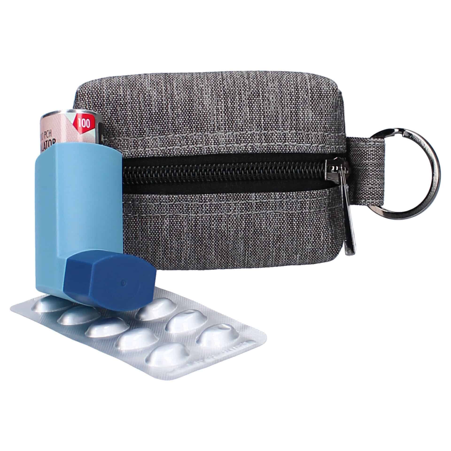 Graue Stofftasche mit Reißverschluss und Schlüsselring, daneben ein blauer Inhalator und eine Blisterpackung mit Pillen.