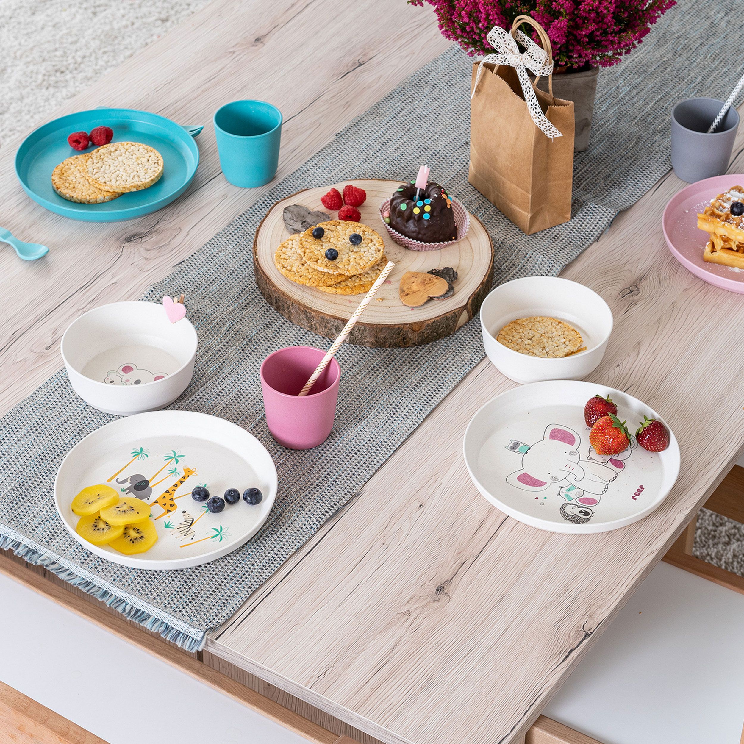 Kindergeschirr-Set auf einem Tisch. Teller mit Wildtier-Design, Beeren und Kuchen. Rosa und blaue Becher. Geschenkpapier.