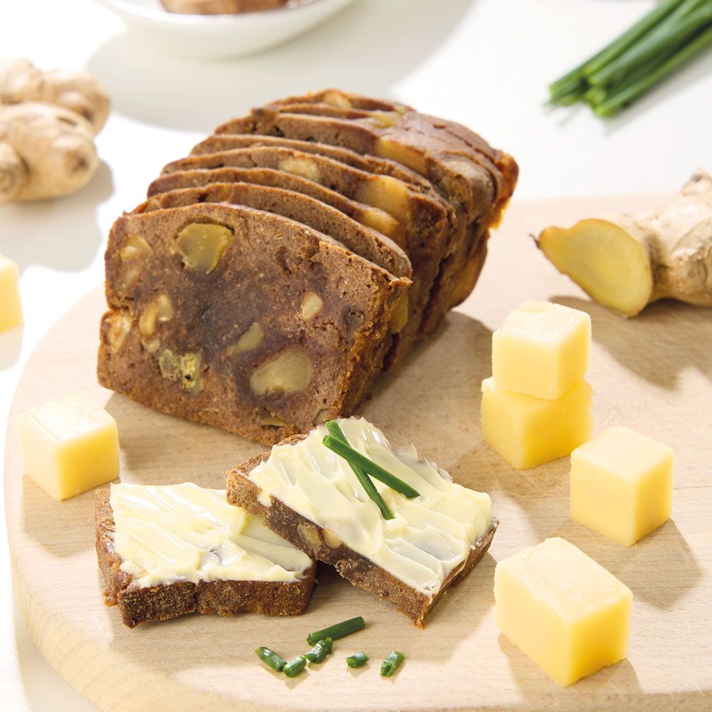 Geschnittenes Kletzzzbrot Ingwer auf Holzbrett. Neben Käsewürfeln und Ingwerknolle. Zwei Brotscheiben mit Belag.