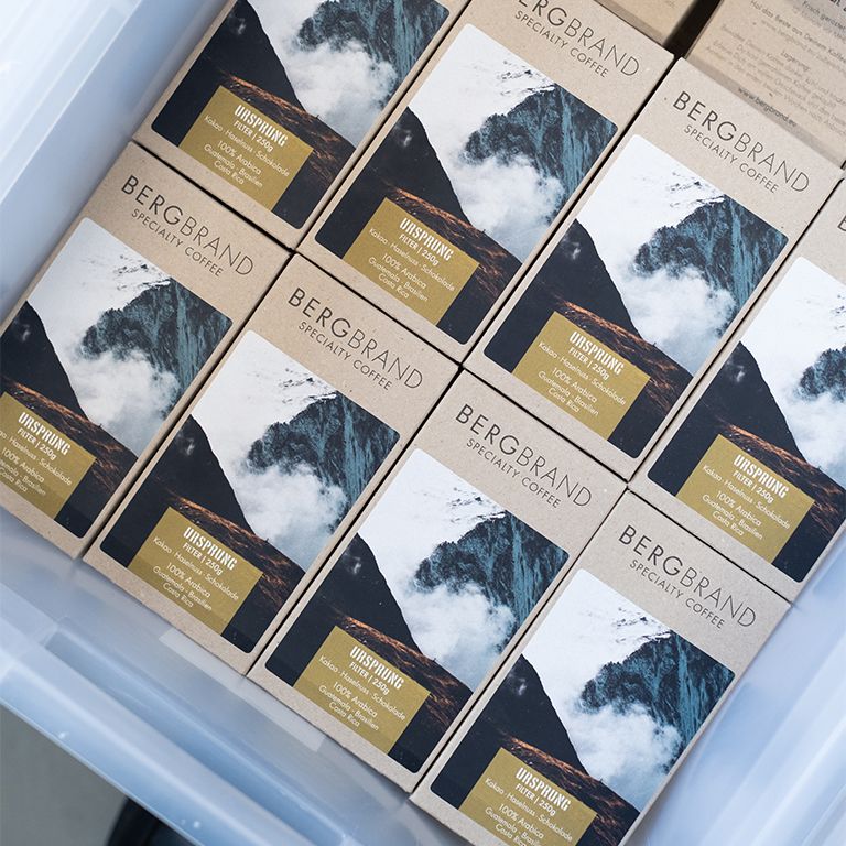 Kaffeepackungen in einer Kiste. Text: BERGBRAND, Ursprung, Filter, 250g. Zutaten: Kakao, Haselnuss, Schokolade, 100% Arabica.