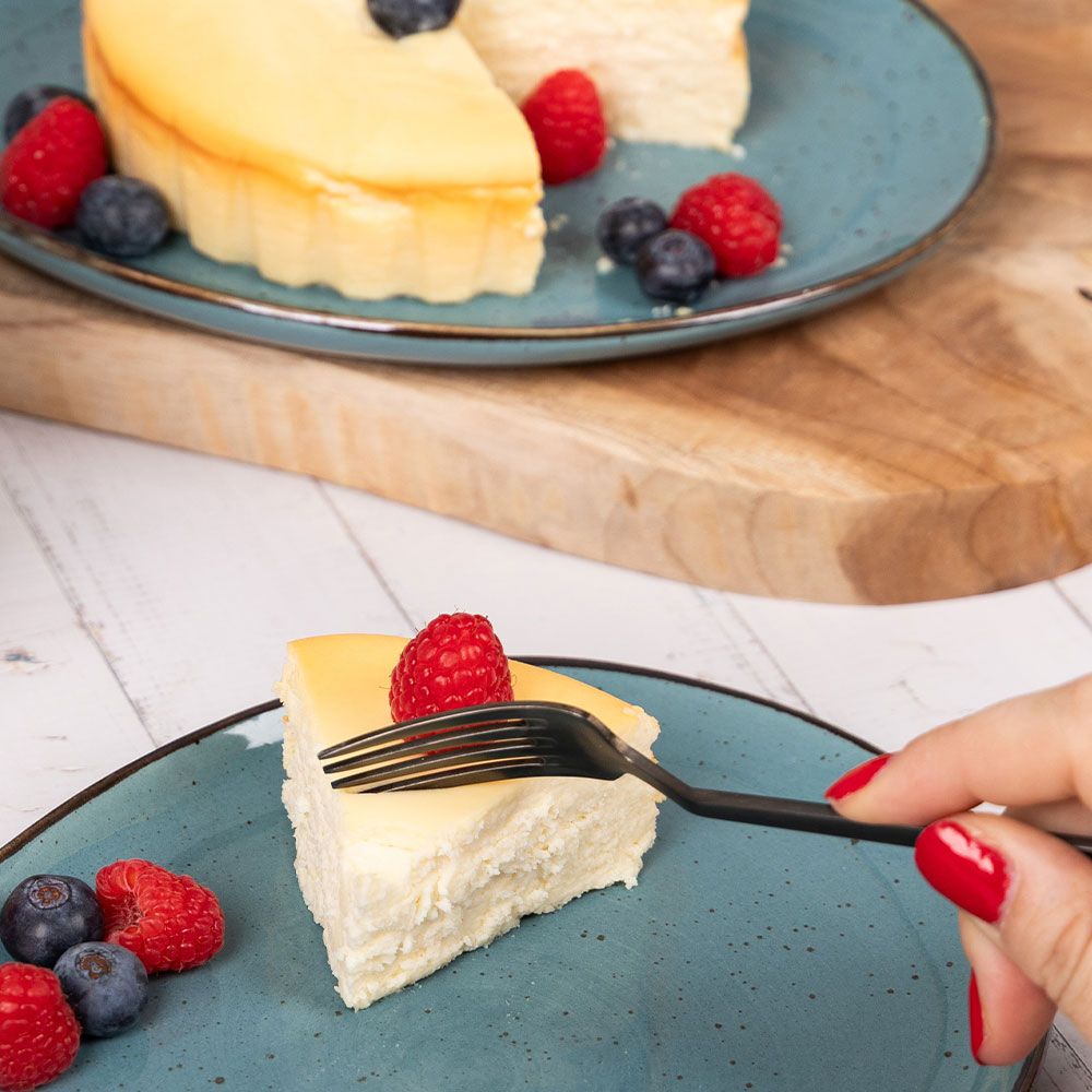 Ein Stück Quarkkuchen auf einem blauen Teller mit Beeren. Eine Hand mit rotem Nagellack hält eine Gabel.