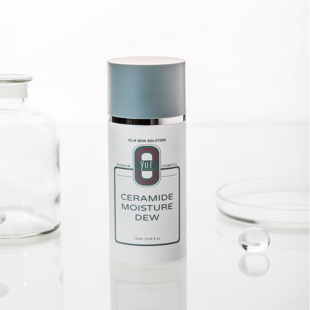 Weiße Lotion-Flasche mit blauem Deckel. Aufschrift: Ceramide Moisture Dew. Marke: Yu.R Skin Solution. Im Hintergrund Glaswaren.