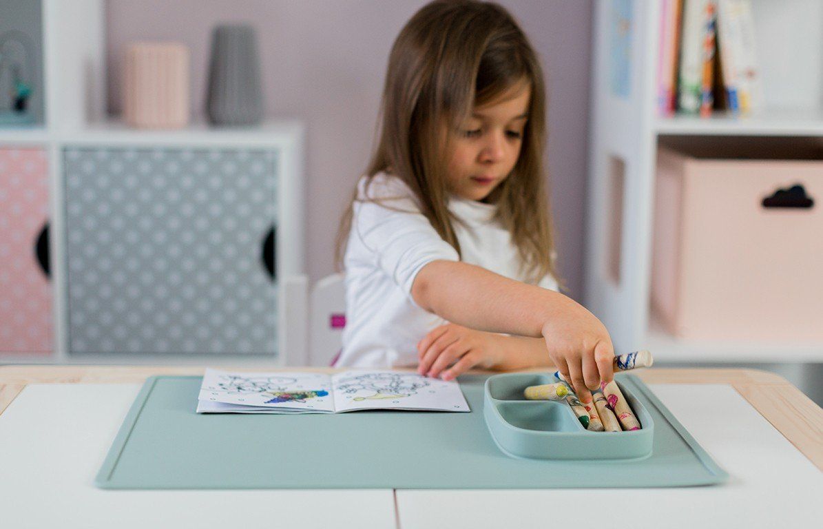 Kind sitzt an Tisch, malt auf Papier. Aquamarinfarbene Spielmatte mit integriertem Teller. Buntstifte im Teller.