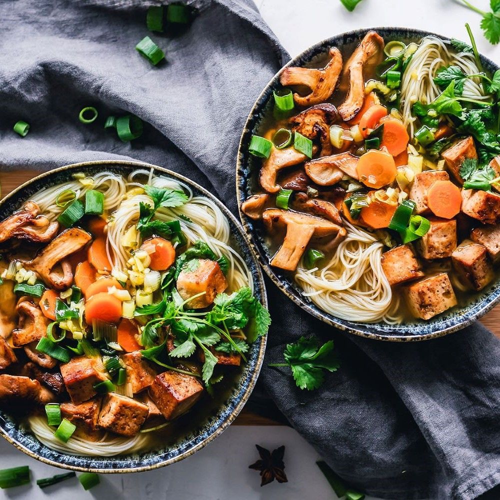 Zwei Schalen Pho-Suppe mit Nudeln, Tofu, Pilzen, Karotten und Kräutern. Auf grauem Tuch. Draufsicht.