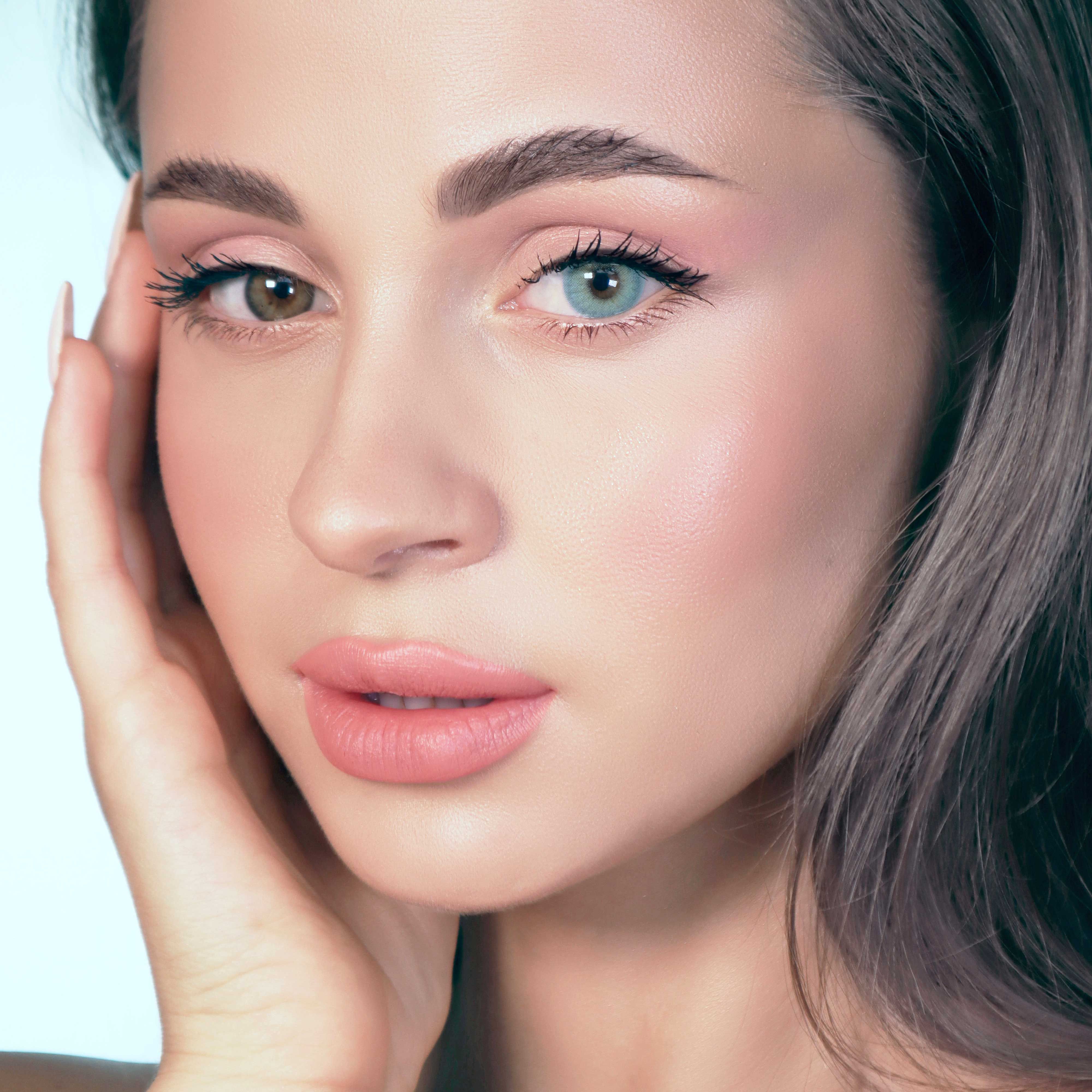 Gesicht einer Frau mit zwei unterschiedlich farbigen Augen. Ein Auge ist braun, das andere türkisblau. Die Frau berührt ihr Gesicht.