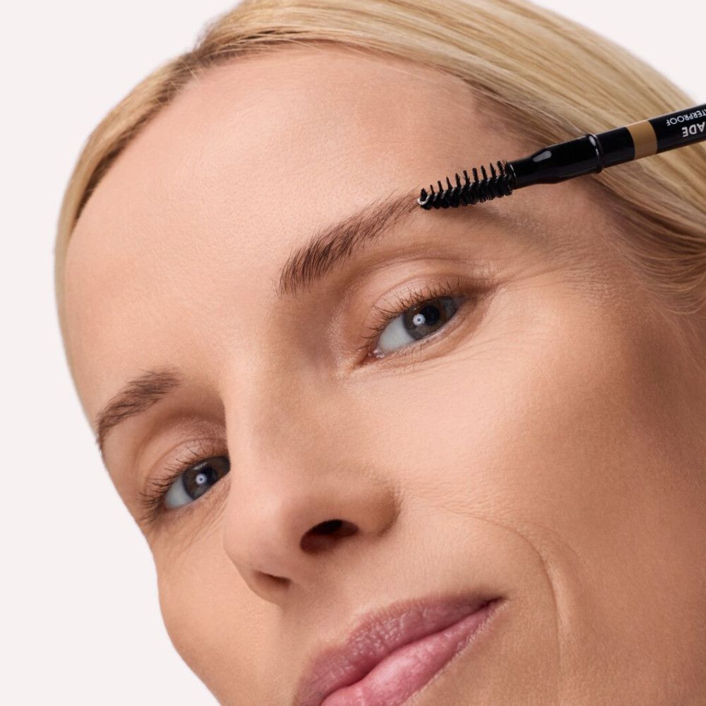 Frau mit Augenbrauenstift. Stift wird an Augenbraue angesetzt. Bürste am Stiftende. Marke: Catrice, Aufschrift: Shape & Shade.
