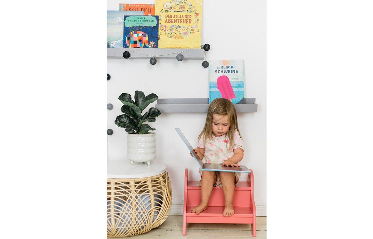 Kind sitzt auf einem Holztritt, altrosa, zweistufig. Bücherregal im Hintergrund.
