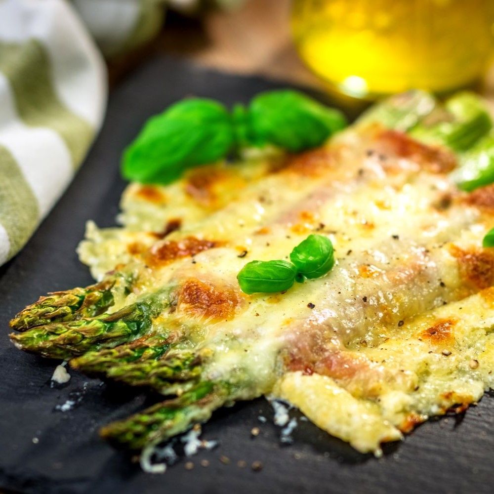 Spargelgericht mit Käse überbacken, auf schwarzem Brett. Basilikumblätter als Garnitur.