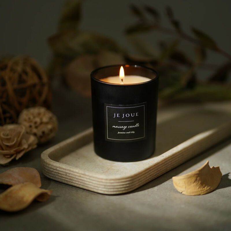 Schwarze Kerze mit Flamme auf einem Tablett. Aufschrift: Je Joue, massage candle, Jasmine Lily. Dekoration im Hintergrund.