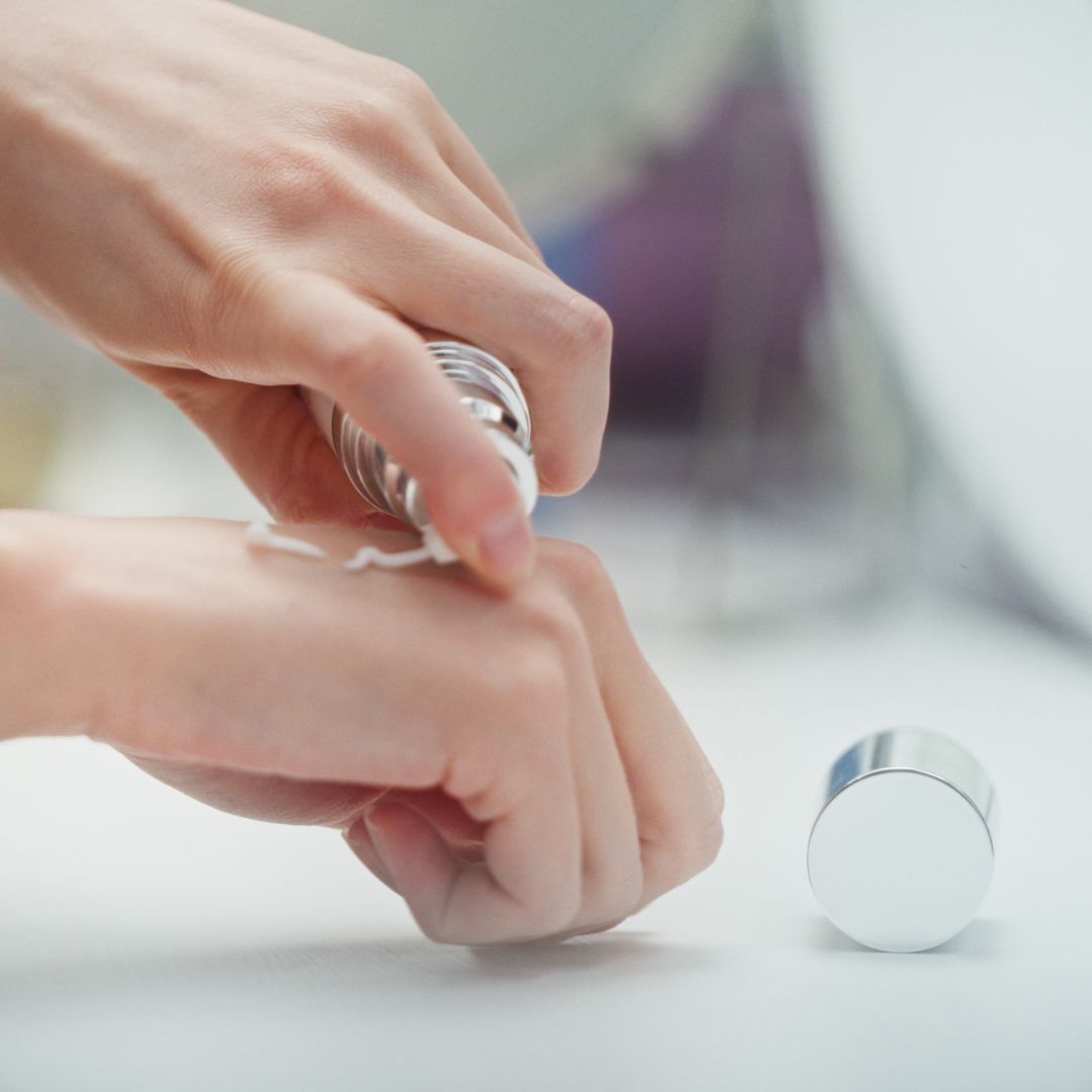 Hände tragen Creme auf. Flasche und Deckel liegen daneben. Creme wird auf die Haut aufgetragen.