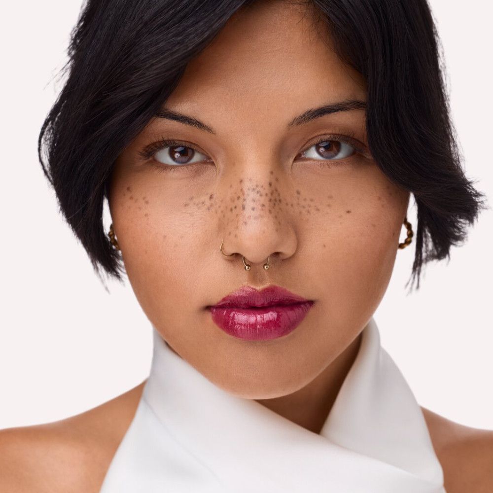 Porträt einer Frau mit aufgetragenem Lipgloss. Sie trägt ein weißes Oberteil und hat einen Nasenring.