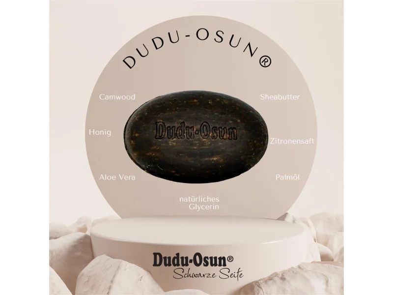 Dudu-Osun Seife, oval, dunkelbraun, auf Podest. Aufschrift 'Dudu-Osun Schwarze Seife'. Zutaten-Labels.