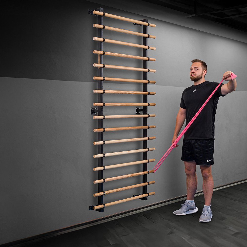 Mann mit Theraband an Sprossenwand. Er steht und trainiert. Schwarzer Rahmen, Holzsprossen.
