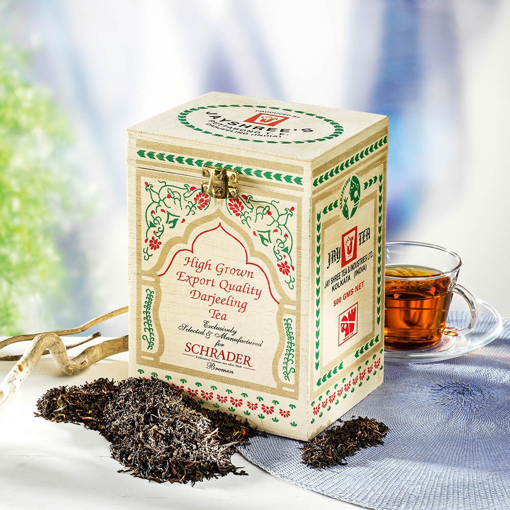 Holzkiste mit "High Grown Export Quality Darjeeling Tea" für Schrader, daneben eine Tasse Tee und lose Teeblätter.