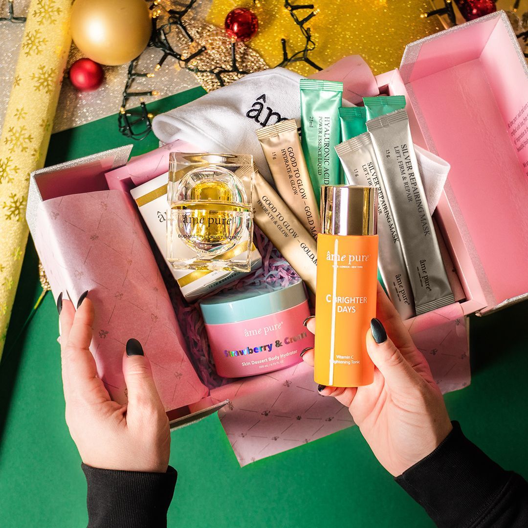 Geschenkbox mit Kosmetikprodukten. Enthält Cremes, Masken und eine orangefarbene Flasche. Gehalten von Händen auf grünem Hintergrund.