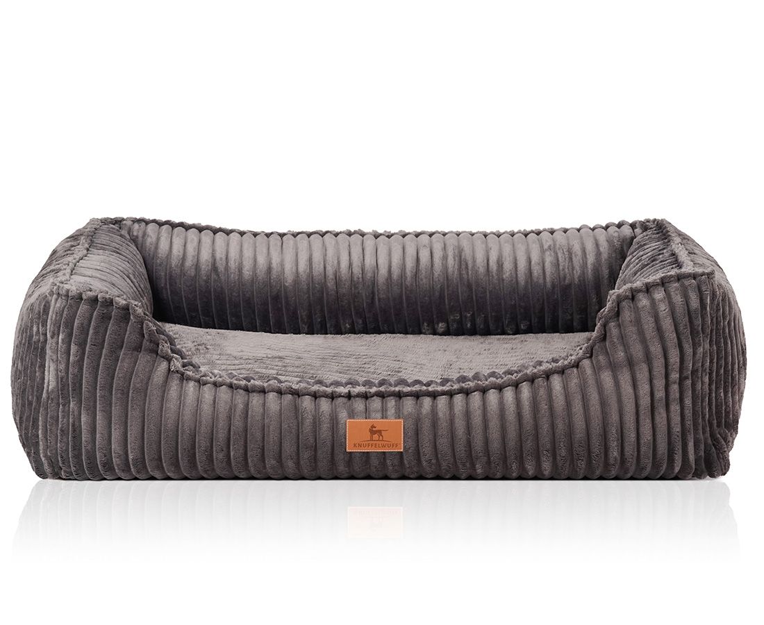Graues, rechteckiges Hundebett mit erhöhten Seiten und geriffelter Oberfläche. Ein braunes Label mit Logo ist angebracht.