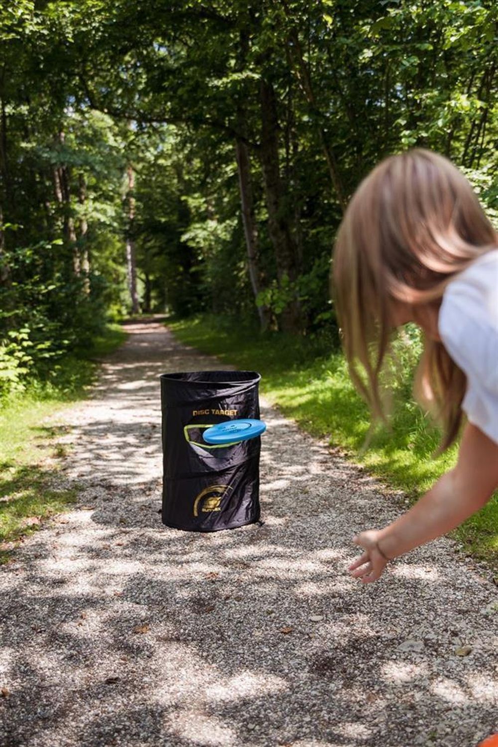 Person wirft blaue Disc Golf Scheibe (Putter) in Richtung eines schwarzen Ziels. Kiesweg im Wald.