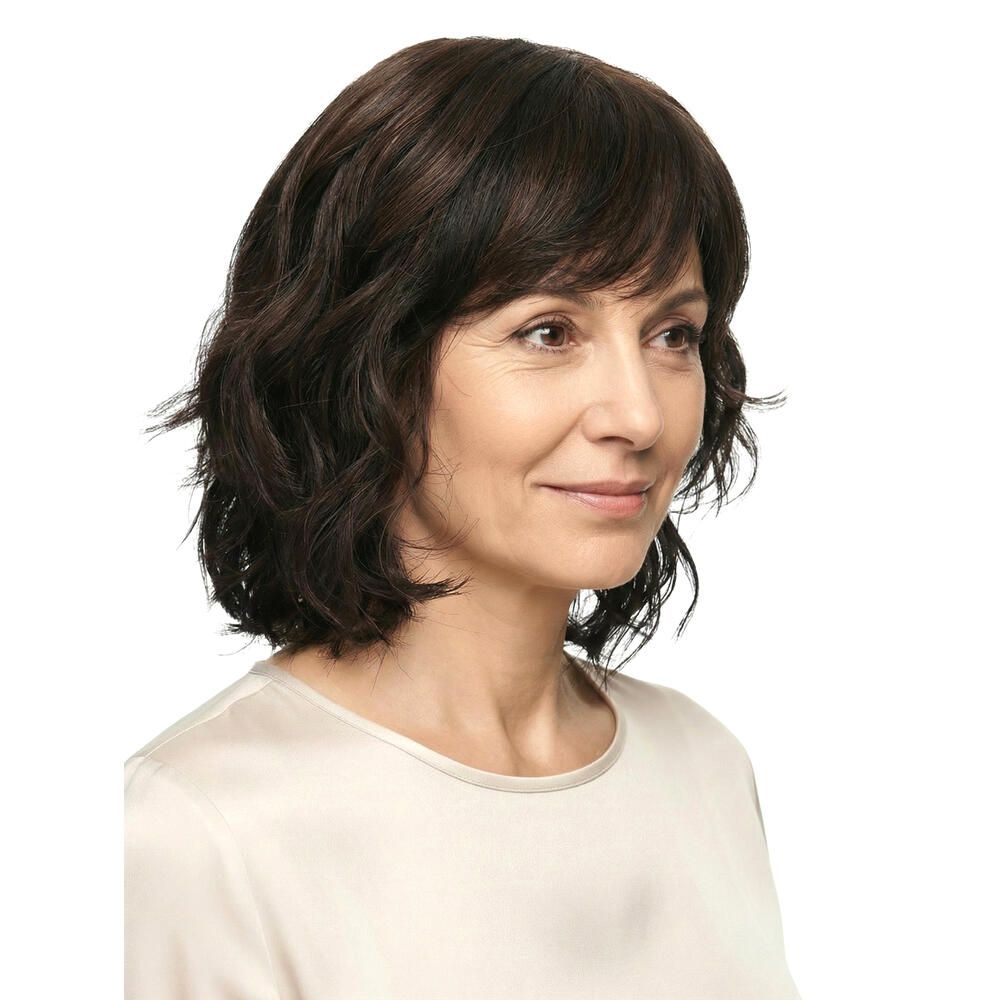 Perücke Capri, braun, gewellt, mit Pony. Frau im Profil, Blick nach rechts. Helles Oberteil.