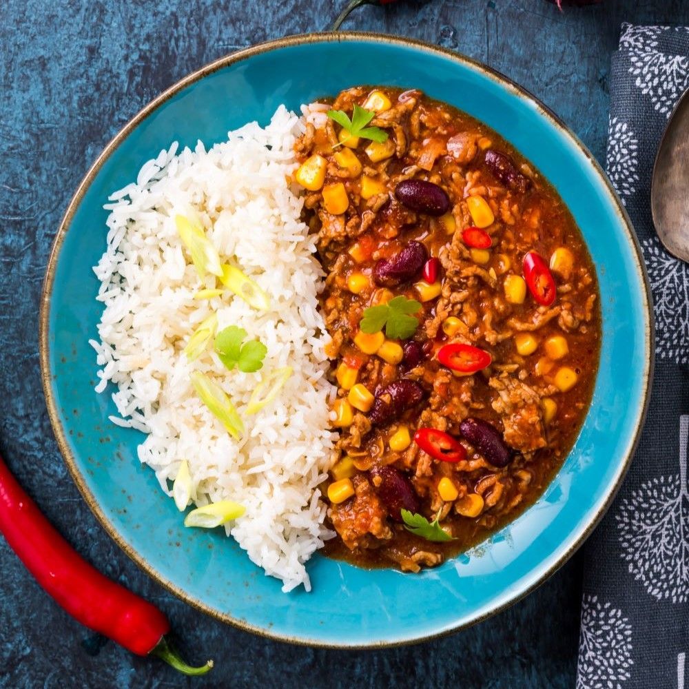 Teller mit Chili con Carne und Reis. Chili mit Mais und Bohnen. Roter Chili am Rand. Blaue Platte.