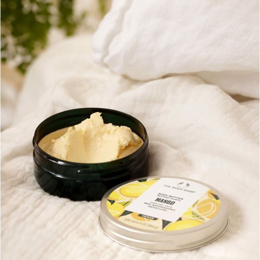 Geöffnete Dose Body Butter Mango, daneben Deckel. Auf weißem Stoff. Aufschrift: Body Butter Mango, für trockene Haut, 96h Feuchtigkeit.