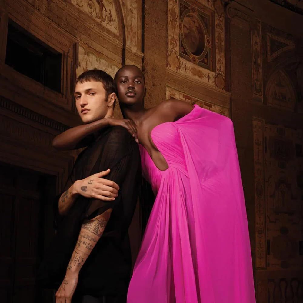 Zwei Personen vor einem Wandgemälde. Eine Person trägt Schwarz, die andere ein pinkfarbenes Kleid.