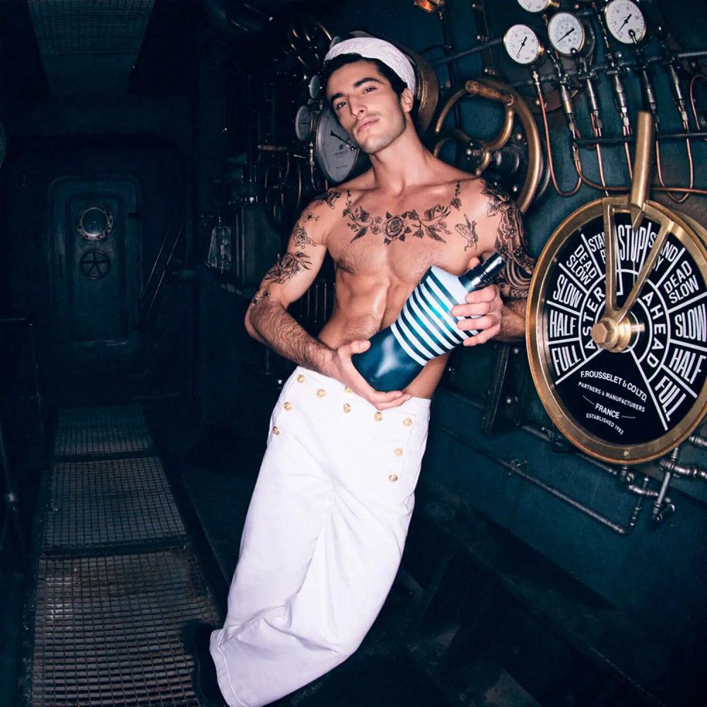 Mann mit Tattoo und Matrosenkleidung hält einen Flakon. Im Hintergrund: Steuerrad und Instrumente.