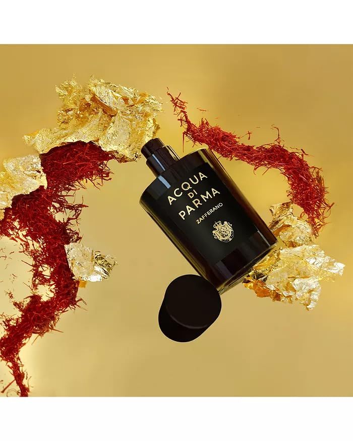 Schwarzer Flakon mit Aufschrift ACQUA DI PARMA Zafferano. Offener Deckel daneben. Auf goldenem Hintergrund mit Safran und Blattgold.