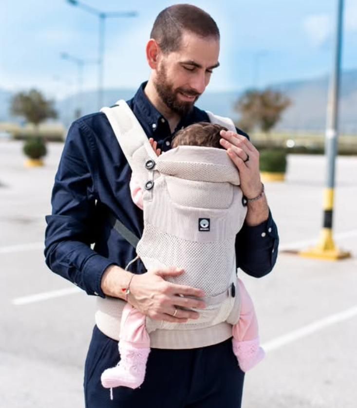 Ein Mann trägt ein Baby in einer beige Babytrage. Das Baby ist nach innen gerichtet.