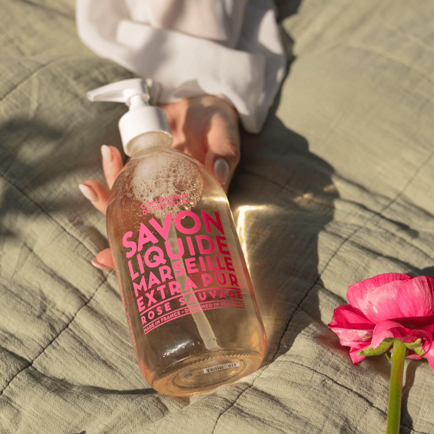 Eine Hand hält eine transparente Flasche mit rosa Etikett. Aufschrift: Savon Liquide Marseille Extra Pur Rose Sauvage. Daneben eine Rose.