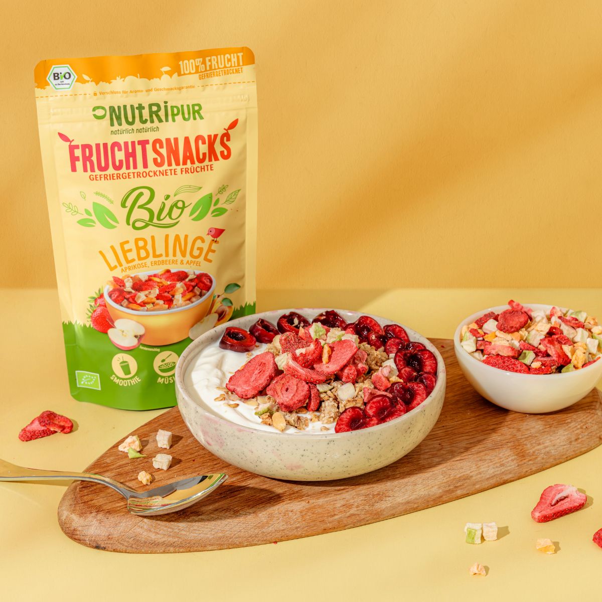 Schale mit Joghurt und gefriergetrockneten Früchten. Daneben eine Tüte mit Früchten. Auf einem Holzbrett.