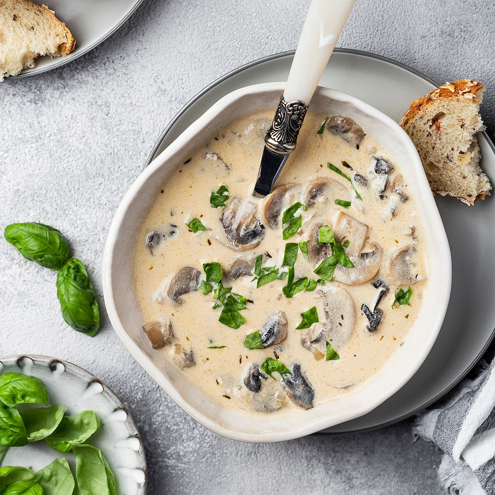 Cremige Suppe mit Pilzen in einer weißen Schale. Garniert mit Kräutern. Auf einem Teller Brot und Basilikumblätter.