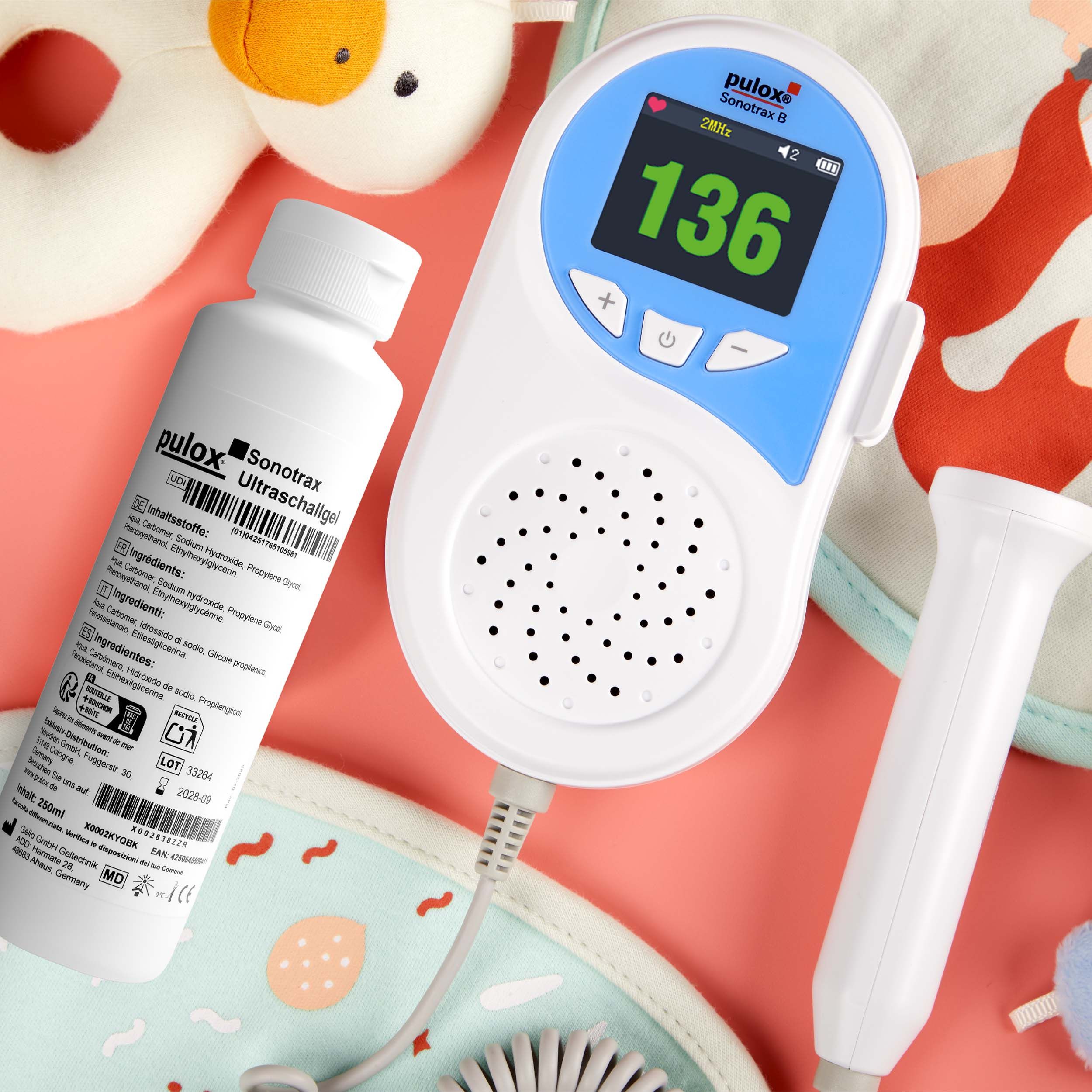 Fetal Doppler und Ultraschallgel auf rosa Untergrund. Doppler zeigt Herzfrequenz 136 an. Mit Spielzeug und Babytuch.