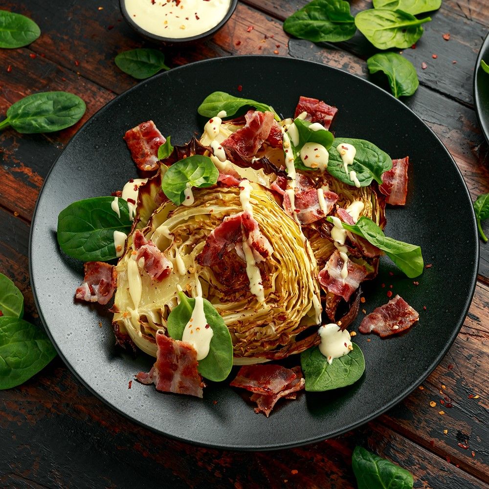 Gebratener Kohlkopf mit Speck und Spinat auf schwarzem Teller. Weißes Dressing. Holzuntergrund.