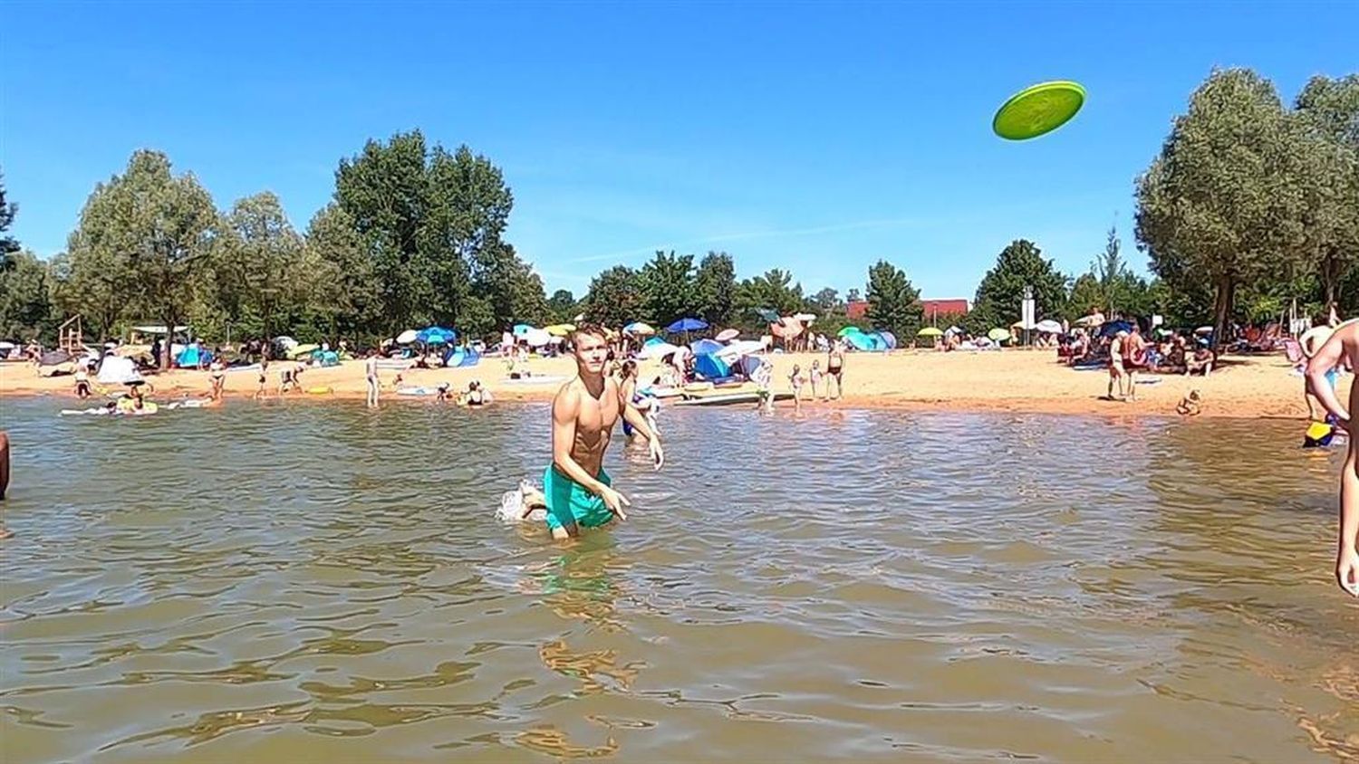 Mann wirft eine grüne Wurfscheibe im Wasser. Am Strand sind weitere Personen zu sehen.