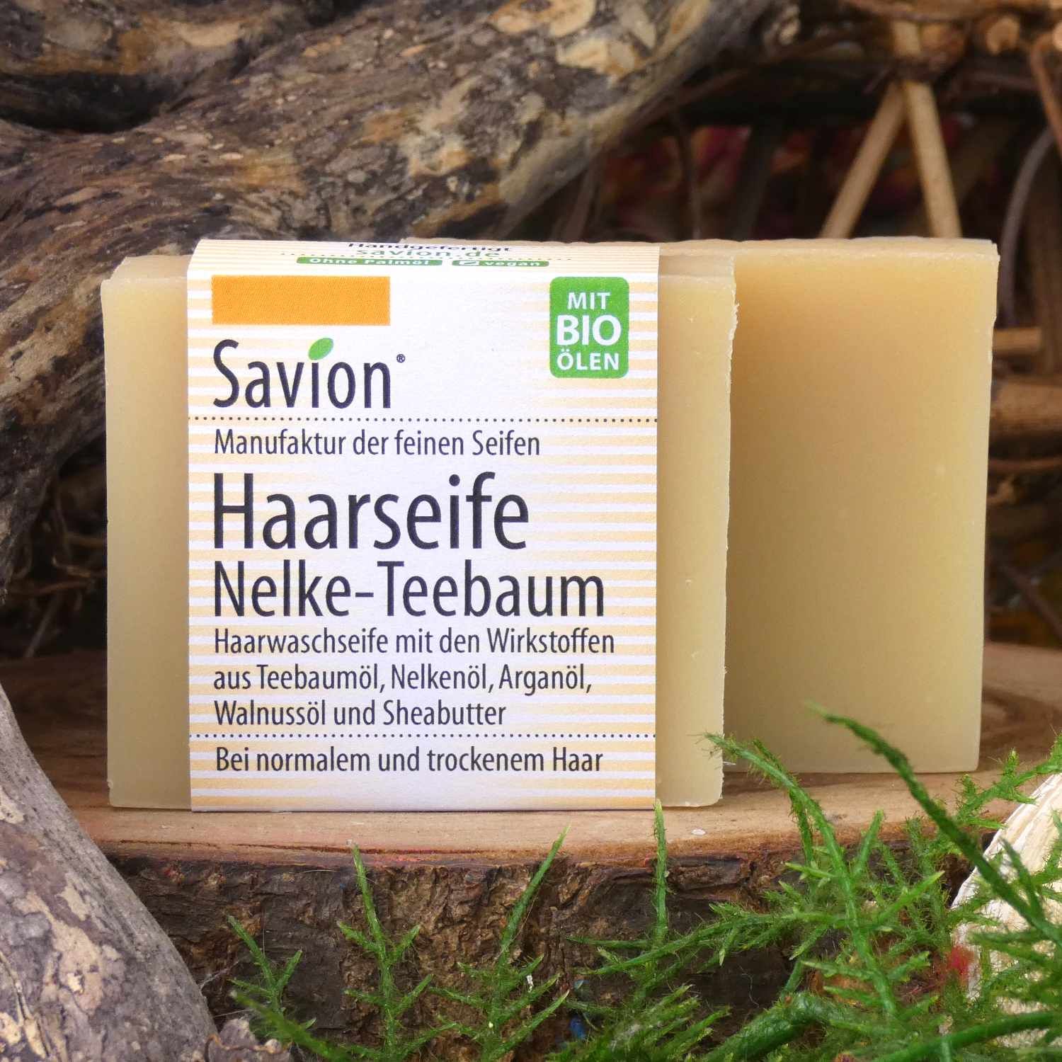 Zwei Seifenstücke auf Holz. Etikett mit Produktinformationen: Haarseife Nelke-Teebaum. Mit Bio-Ölen. Text: Haarwaschseife.