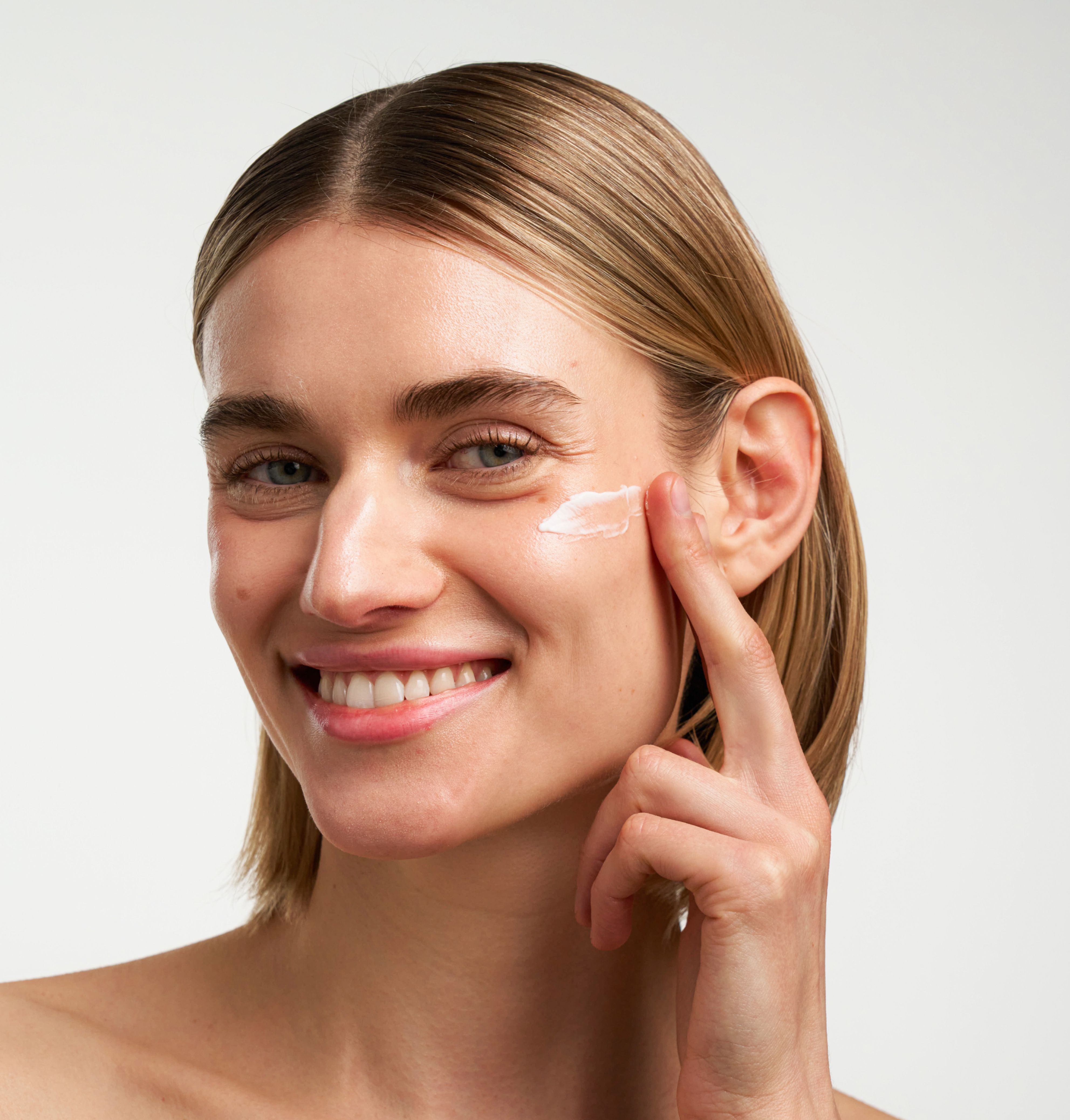 Frau mit aufgetragenem Produkt auf der Haut. Sie lächelt. Helle Haut, blonde Haare.