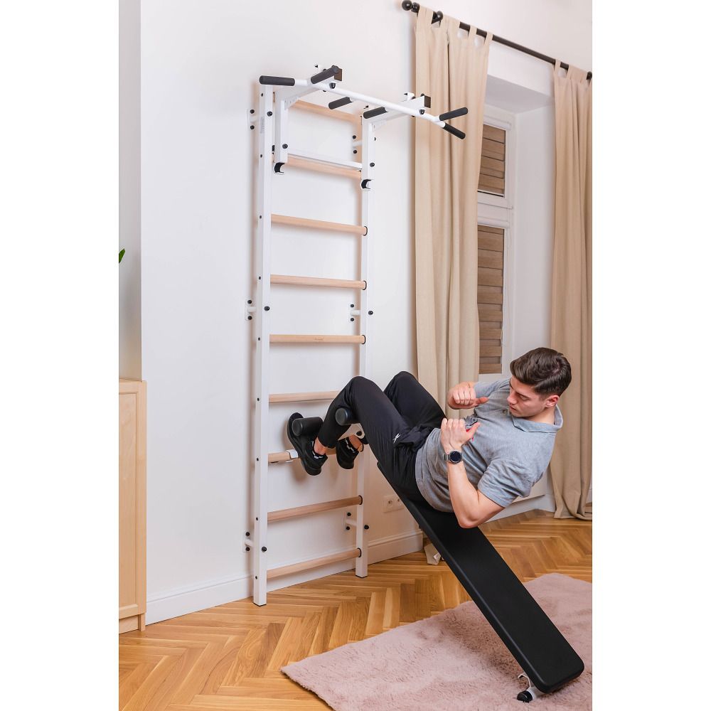 Mann trainiert an einer Sprossenwand mit Trainingsbank. Weiße Sprossenwand, Holzsprossen, schwarze Bank. In einem Raum.