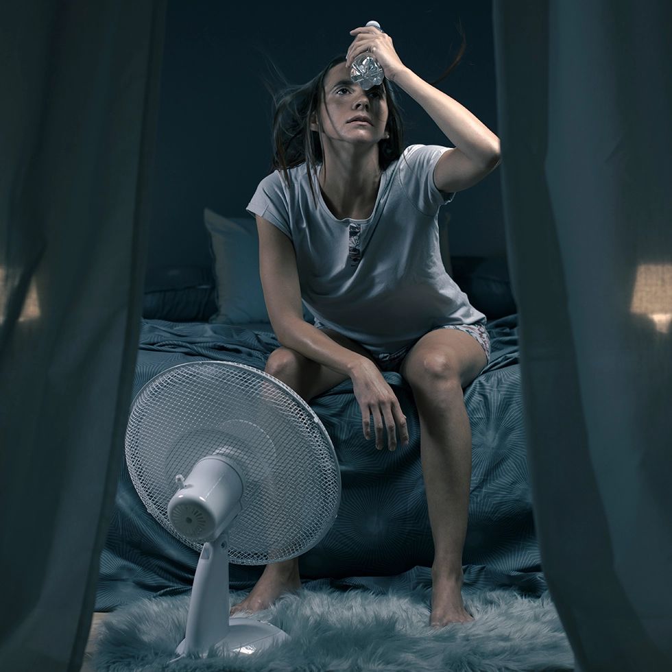 Frau im Bett, hält Wasserflasche an Stirn. Ventilator im Hintergrund.
