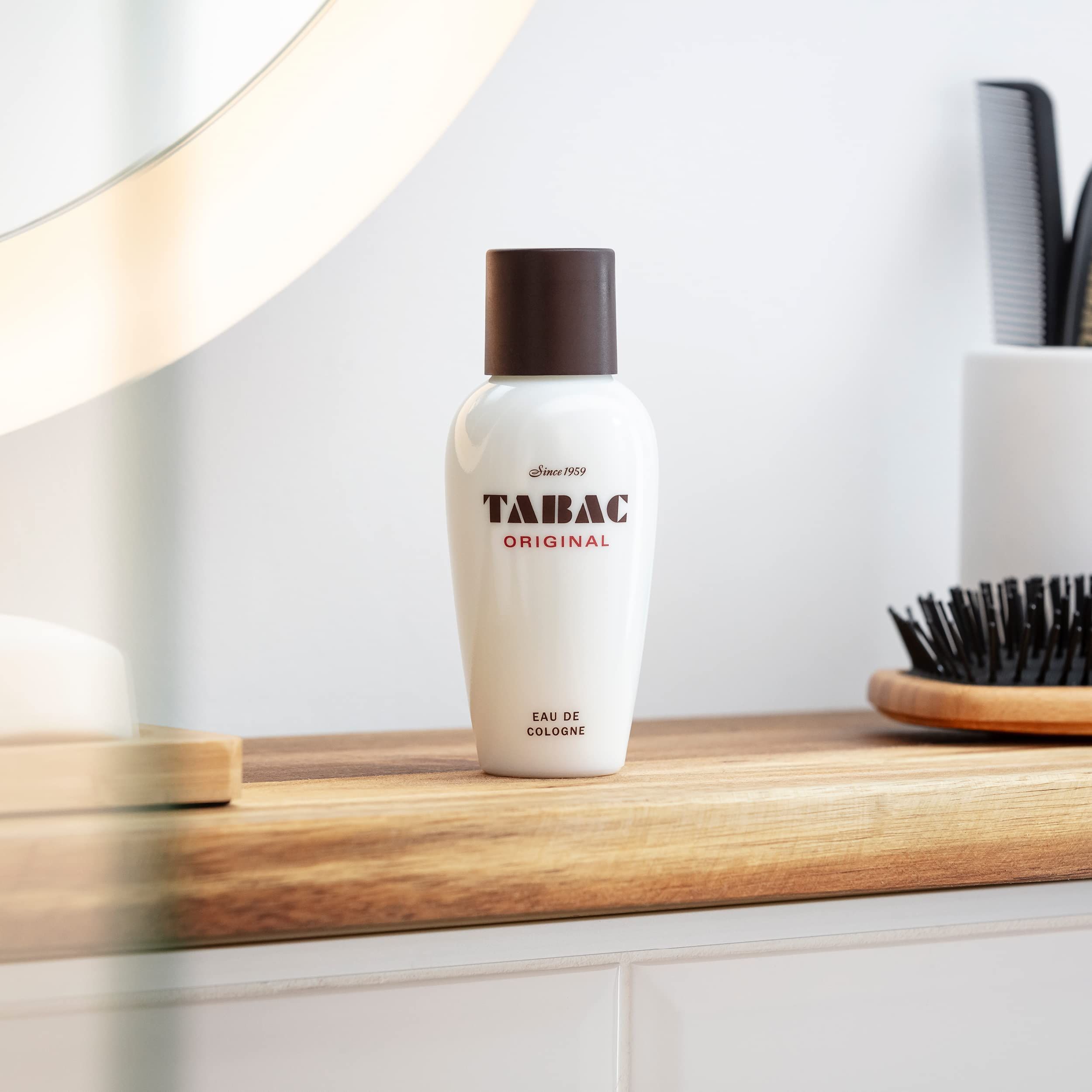 Weiße Flasche mit braunem Deckel. Tabac Original Eau de Cologne. Steht auf Holzablage. Spiegel und Kamm im Hintergrund.