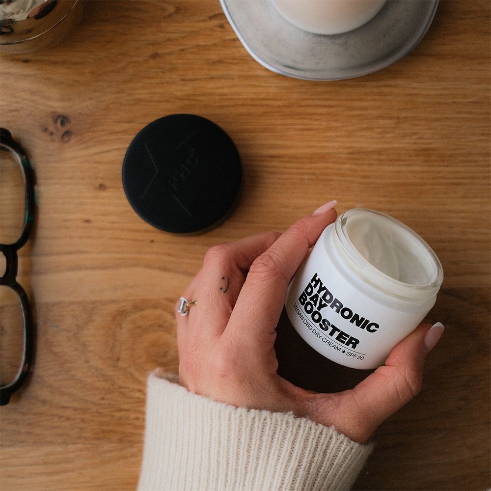 Weißes Tiegel mit schwarzem Deckel. Aufschrift: HYDRONIC DAY BOOSTER, Vegan CBD Day Cream + SPF 20. Gehalten von einer Hand.