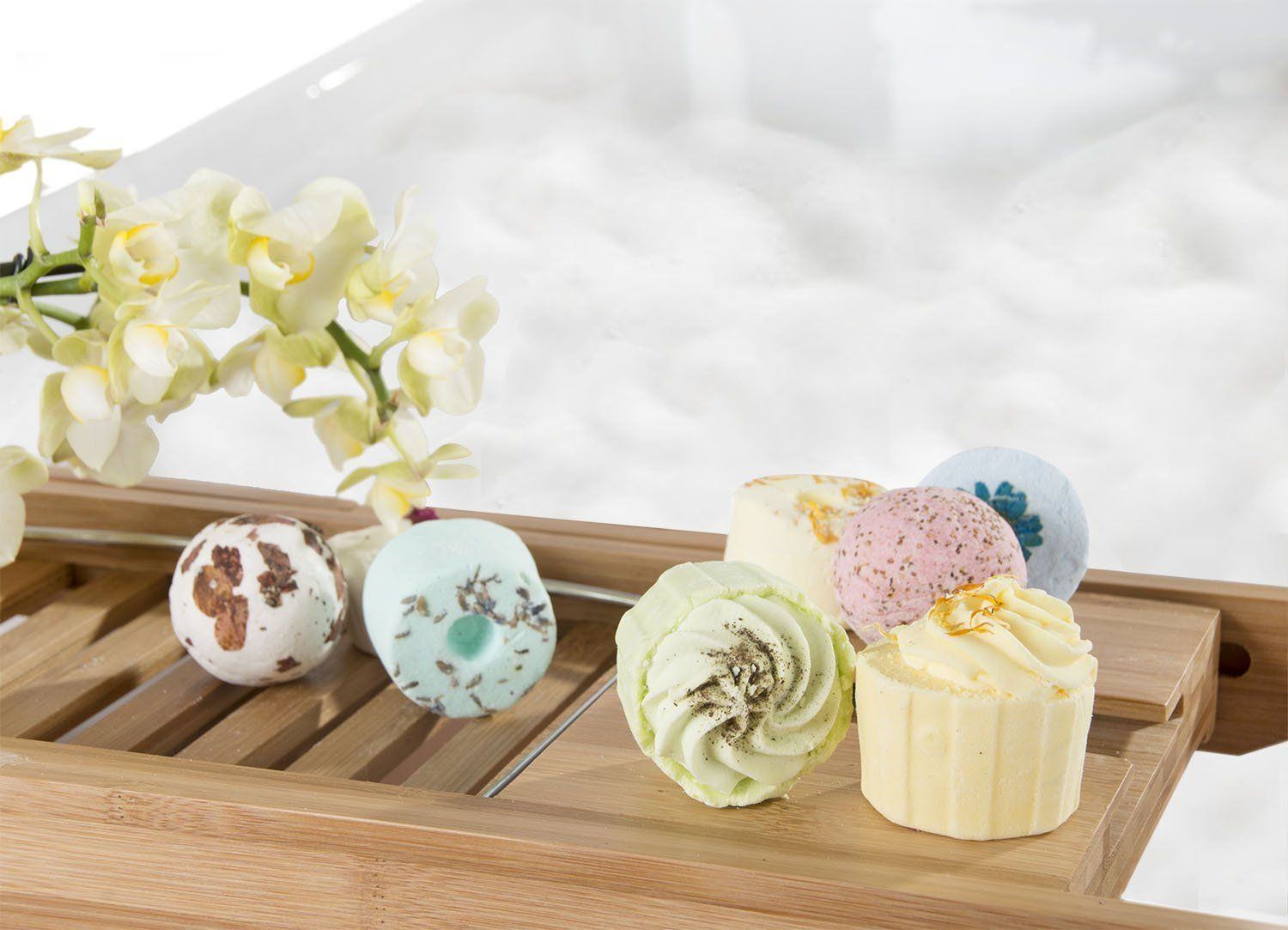 Badebomben und -kuchen auf einem Holztablett, im Hintergrund Schaumbad. Verschiedene Formen und Farben.