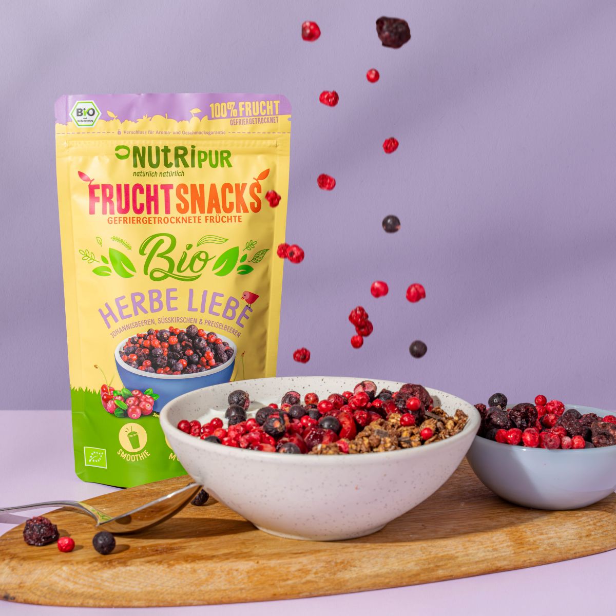 Produktverpackung und Schalen mit gefriergetrockneten Früchten und Joghurt auf Holzbrett. Einige Früchte fallen. Bio-Siegel.