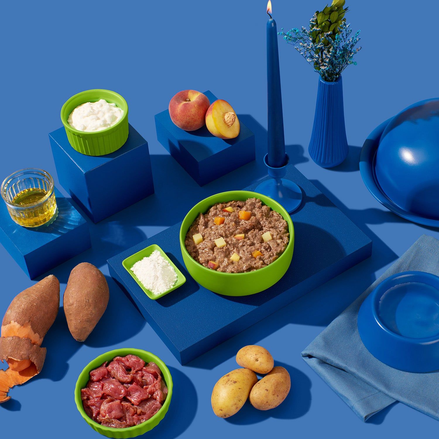 Verschiedene Zutaten auf blauen Podesten. Grüne Schälchen mit Futter, Obst, Gemüse, Öl, Kartoffeln, Kerze, Wassernapf.