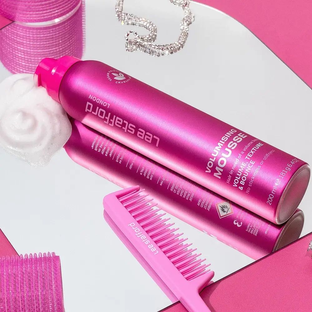 Pinke Dose Lee Stafford Volumising Mousse auf Spiegel, daneben Kamm und Schmuck. Mousse daneben.