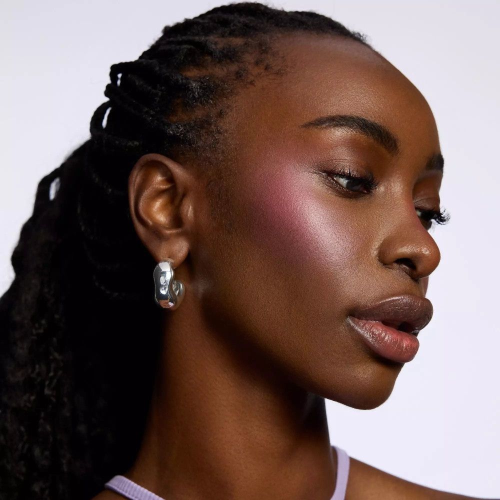 Porträt einer Frau mit rosa Rouge auf den Wangen. Sie trägt silberne Creolen. Ihr Haar ist in Zöpfen geflochten.