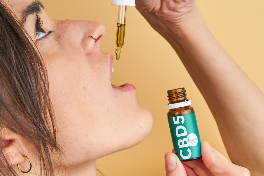 Person öffnet den Mund, um Tropfen aus einer Flasche mit grüner Etikettierung aufzunehmen. Aufschrift CBD5.